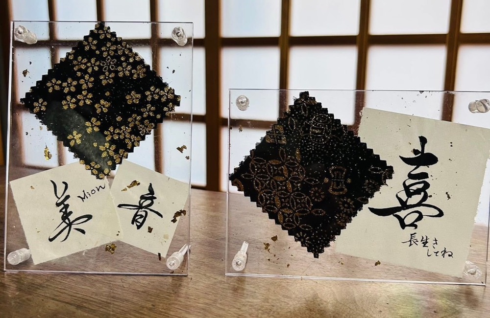 作品の見本 2 点。左は金彩の布と「美音」の書、右はひし形の金彩の布と「喜 長生きしてね」の書。